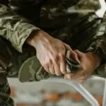 A soldier in camo holding a cap, showing detailed hand gesture and military attire.