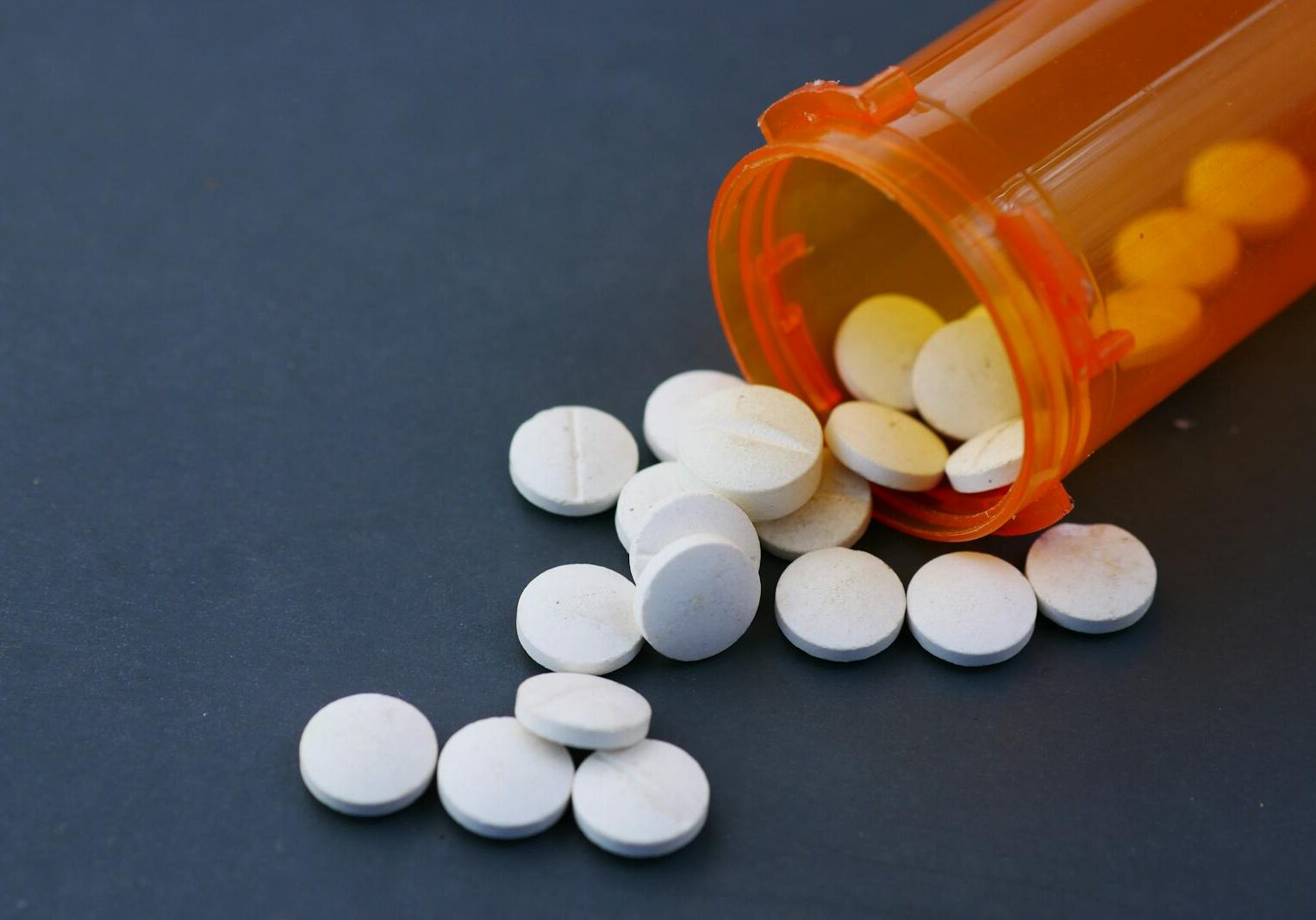 Close-up of white pills scattered from an orange prescription bottle on a gray surface.