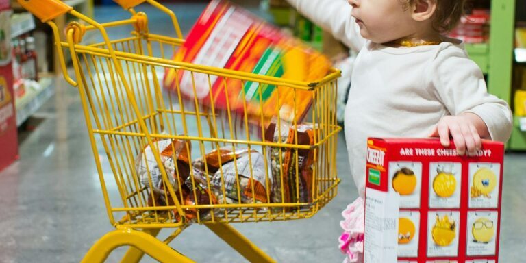 toddle carrying red and white box standing beside yellow shopping cart
