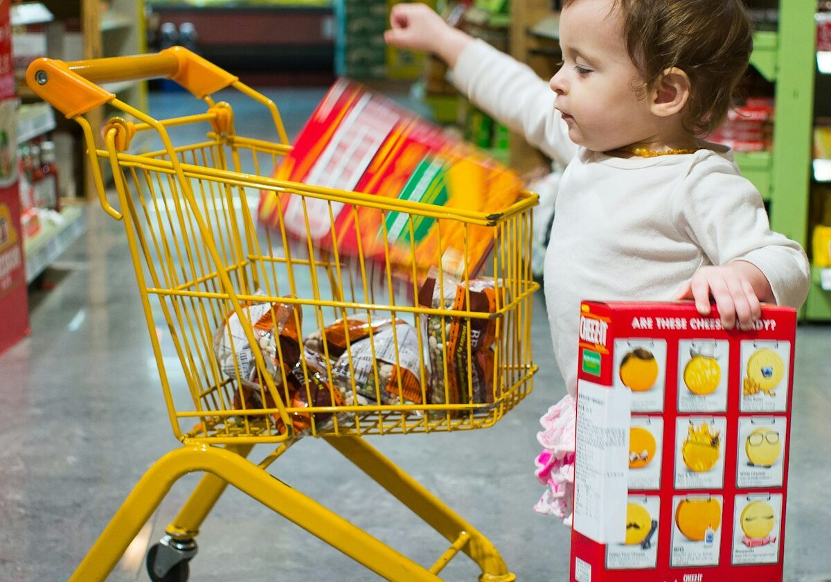 Photo by David Veksler toddle carrying red and white box standing beside yellow shopping cart