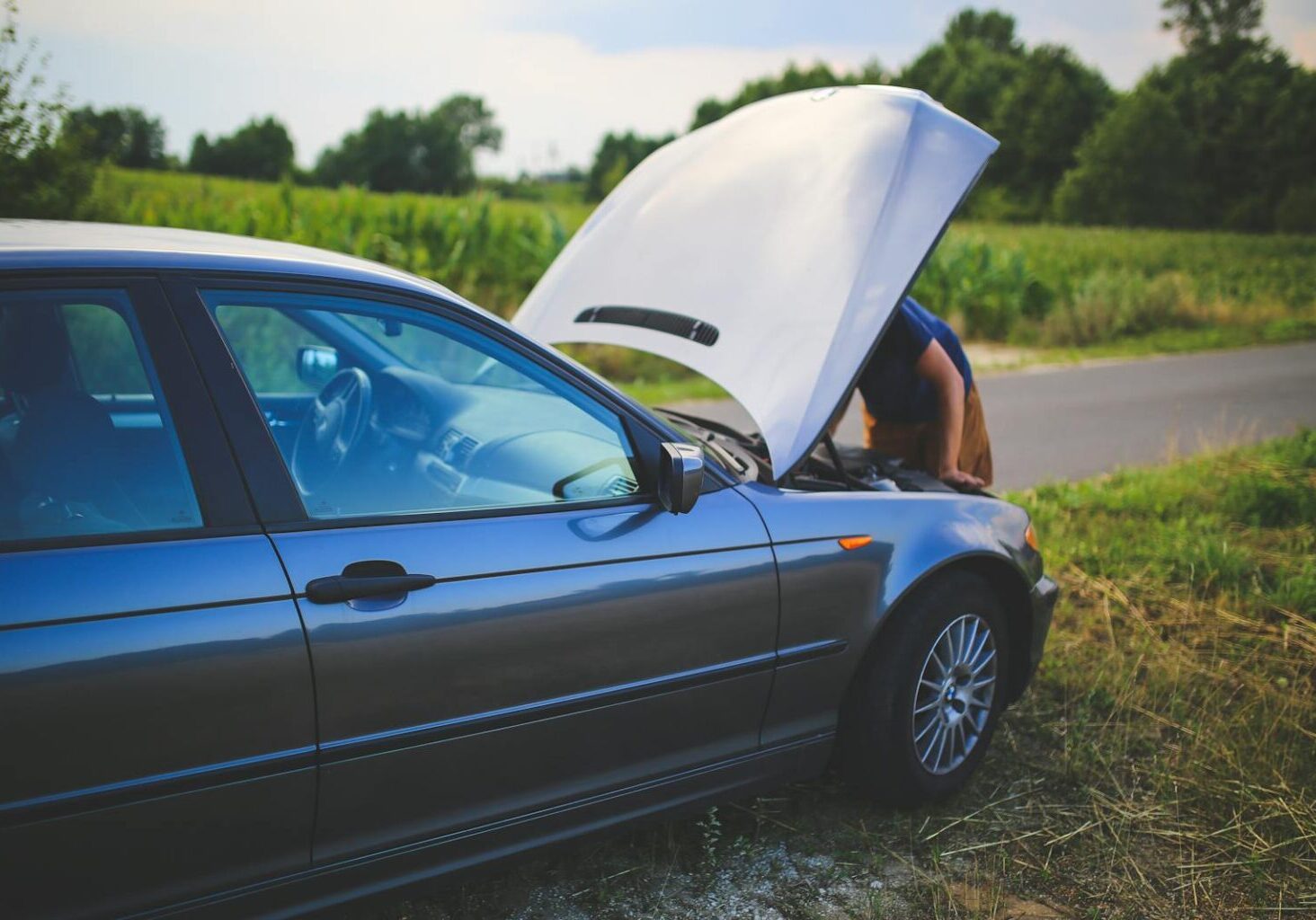 Man checking car engine with hood open by the side of a rural road.