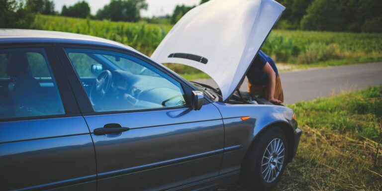 Man checking car engine with hood open by the side of a rural road.