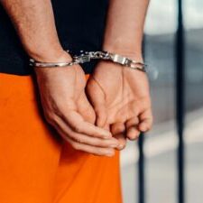Close-up of person in handcuffs wearing orange attire behind prison bars, symbolizing arrest.