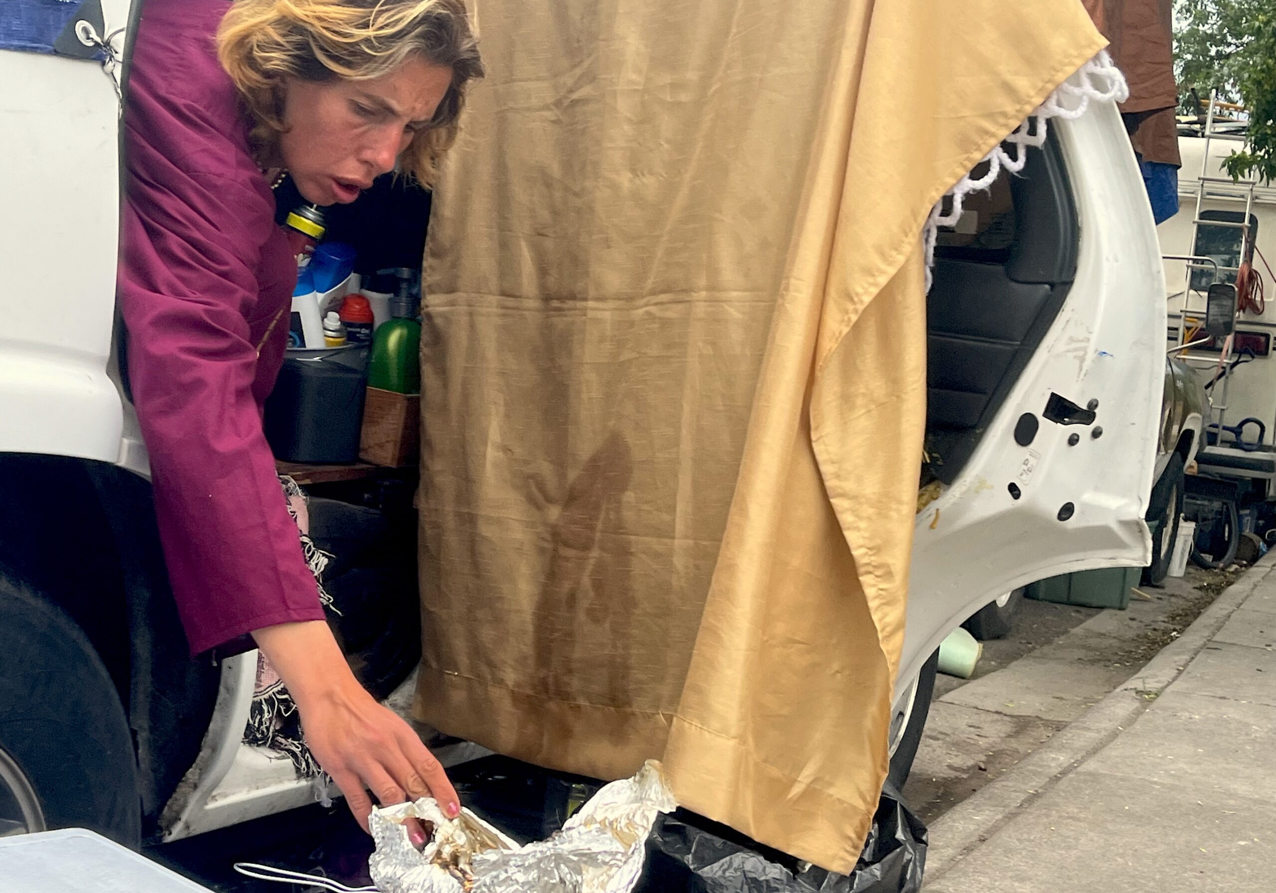 Person leans from the back seat of a car at Hawthorne and Pine behind the Poverello Center to adjust cooking foil on portable camping burner.